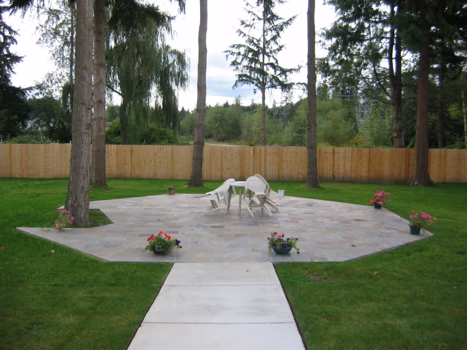 Outdoor patio area with a paved octagonal stone surface surrounded by green grass and trees. There are several potted flowers placed around the patio and a white plastic table with four white plastic chairs, two of which are tipped over. A wooden fence encloses the area and a concrete walkway leads up to the patio.