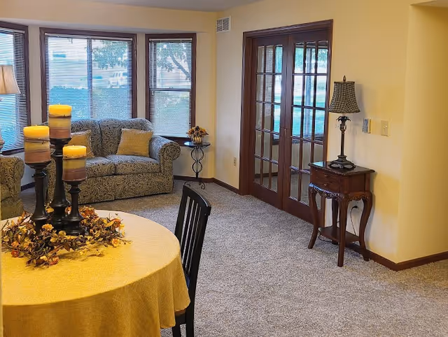 A cozy living area with a round table covered with a yellow tablecloth and decorated with a candle centerpiece. Behind the table is a patterned loveseat with a yellow pillow, positioned in front of three windows with blinds. To the right, there is a wooden side table with a lamp and a set of French doors with glass panes leading to another room. The room has beige walls and carpeted flooring.