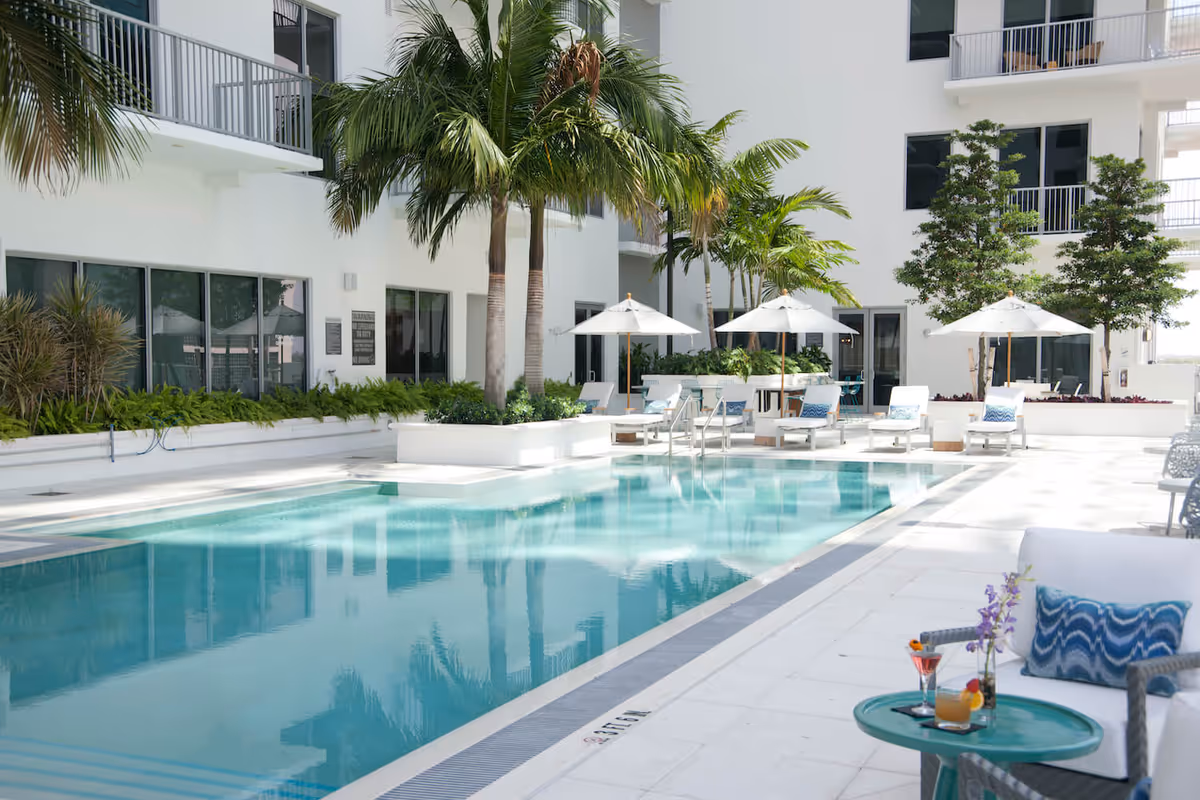 Outdoor swimming pool area at a senior living facility with palm trees, white lounge chairs with blue patterned cushions, white umbrellas, and a small table with drinks and flowers. The building is white with balconies and large windows.