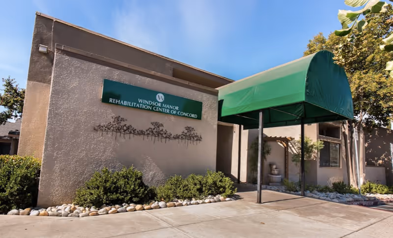 Exterior view of Windsor Manor Rehabilitation Center of Concord showing the building entrance with a green canopy, beige walls, decorative bushes, and a sign with the facility's name on the wall.