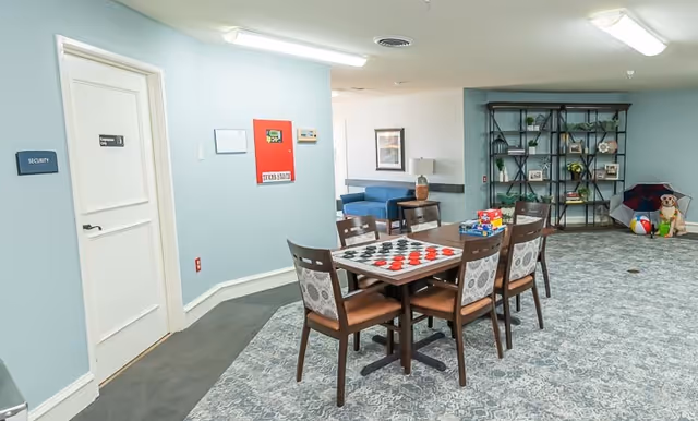 A common area in a senior living facility featuring a table with a checkers board and chairs around it. In the background, there is a blue couch, a bookshelf with decorative items, and a corner with toys including a beach ball and a stuffed dog. The walls are light blue, and there is a door labeled 'Security'.