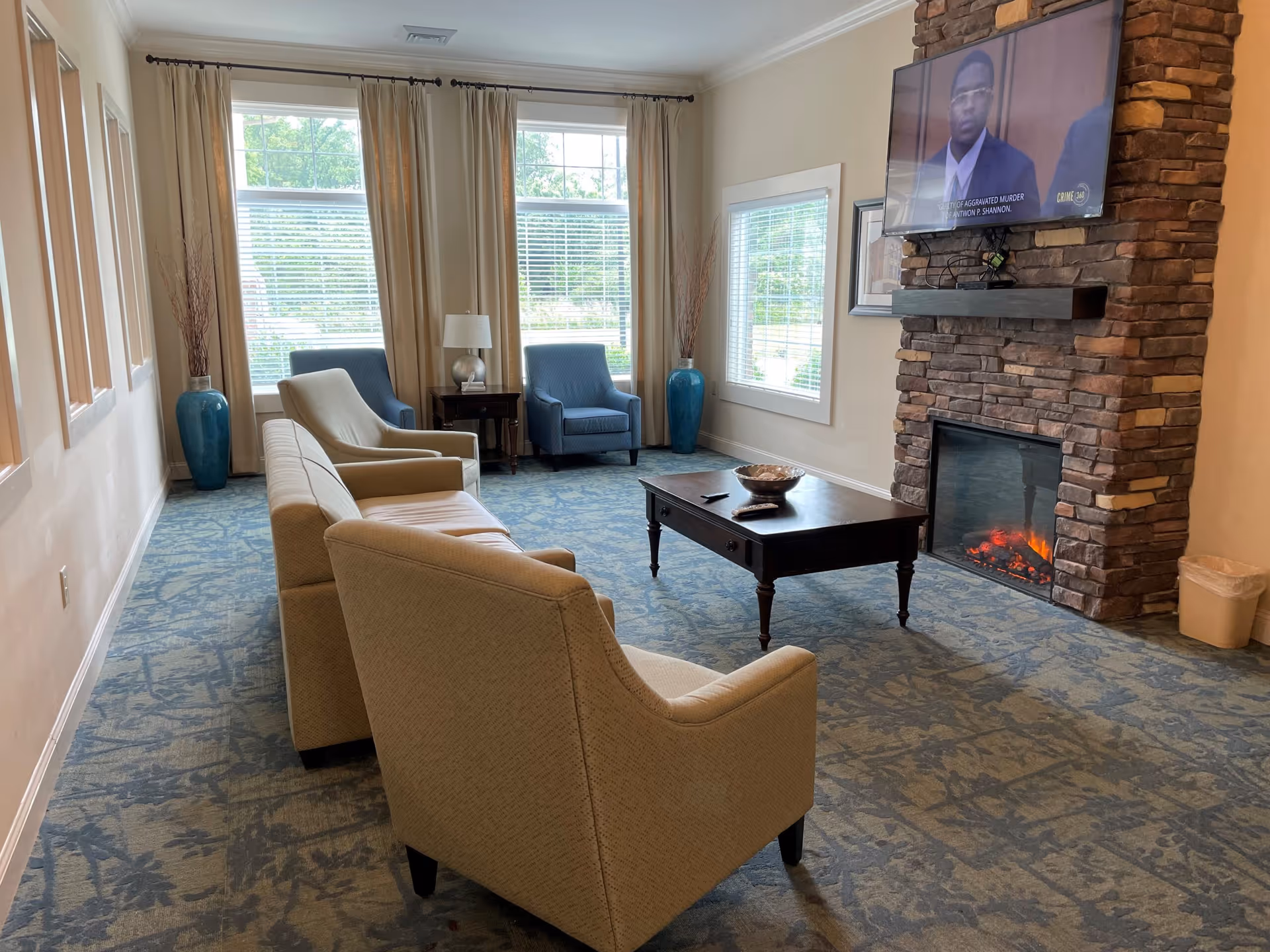 A cozy living room area with a stone fireplace, a mounted flat-screen TV above it, and a coffee table in front. The room has beige walls, large windows with beige curtains, two blue armchairs, and two beige armchairs arranged around the coffee table. There are two tall blue vases with decorative branches near the windows.