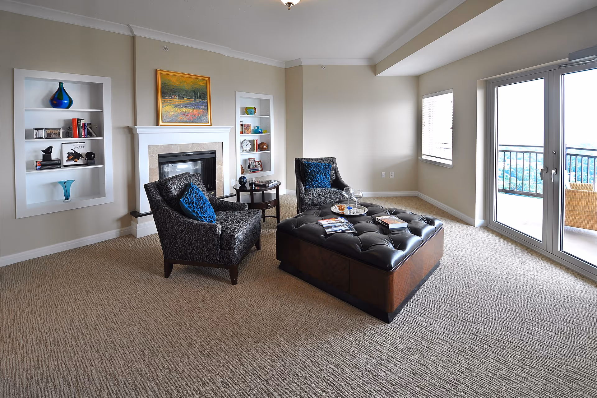 A bright living room with two patterned armchairs with blue cushions, a large tufted leather ottoman with books and a tray on top, a fireplace with a painting above it, built-in shelves with decorative items, and glass doors leading to a balcony with outdoor seating.