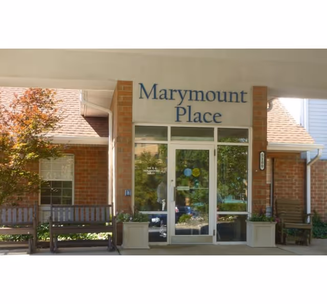 Entrance to Marymount Place Assisted Living facility with glass double doors, brick walls, two wooden benches, potted plants, and a tree with autumn-colored leaves on the left side.