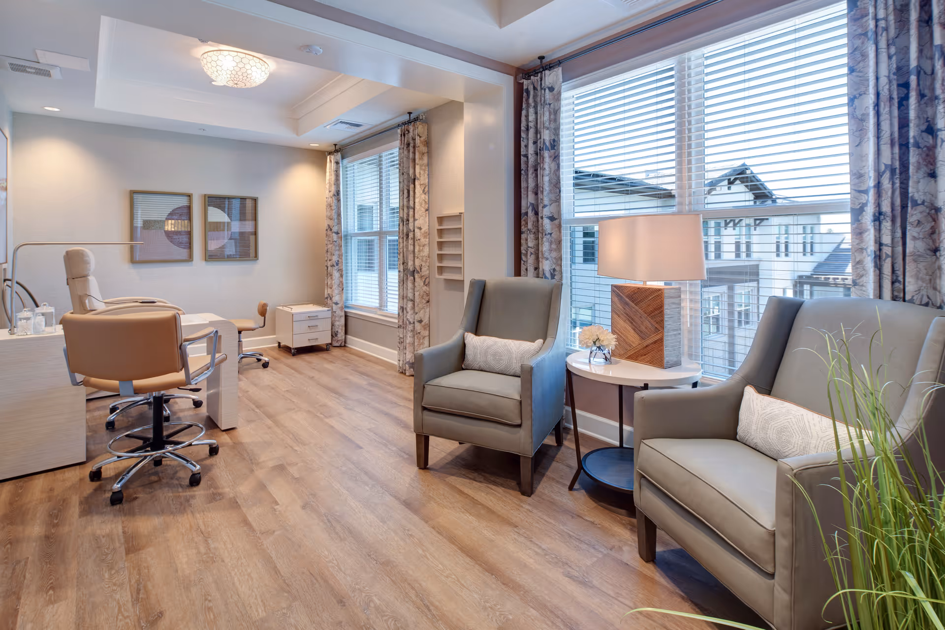 Bright seating area with two armchairs and a side table with a lamp next to large windows, plus a desk and rolling chairs on wood flooring.