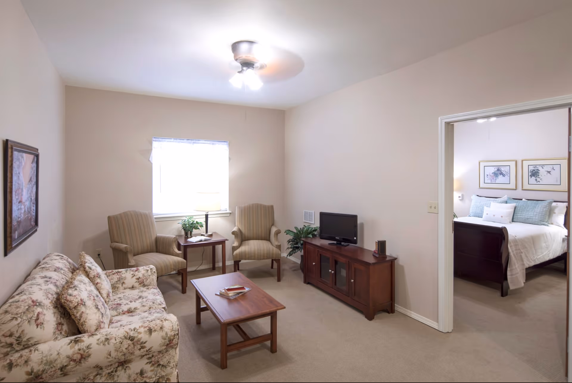 A cozy living room with a floral patterned sofa, two striped armchairs, a wooden coffee table with books, a wooden TV stand with a small television, a side table with a lamp and a plant, and a ceiling fan with lights. Through an open doorway, a bedroom with a bed, pillows, and framed artwork on the wall is visible.