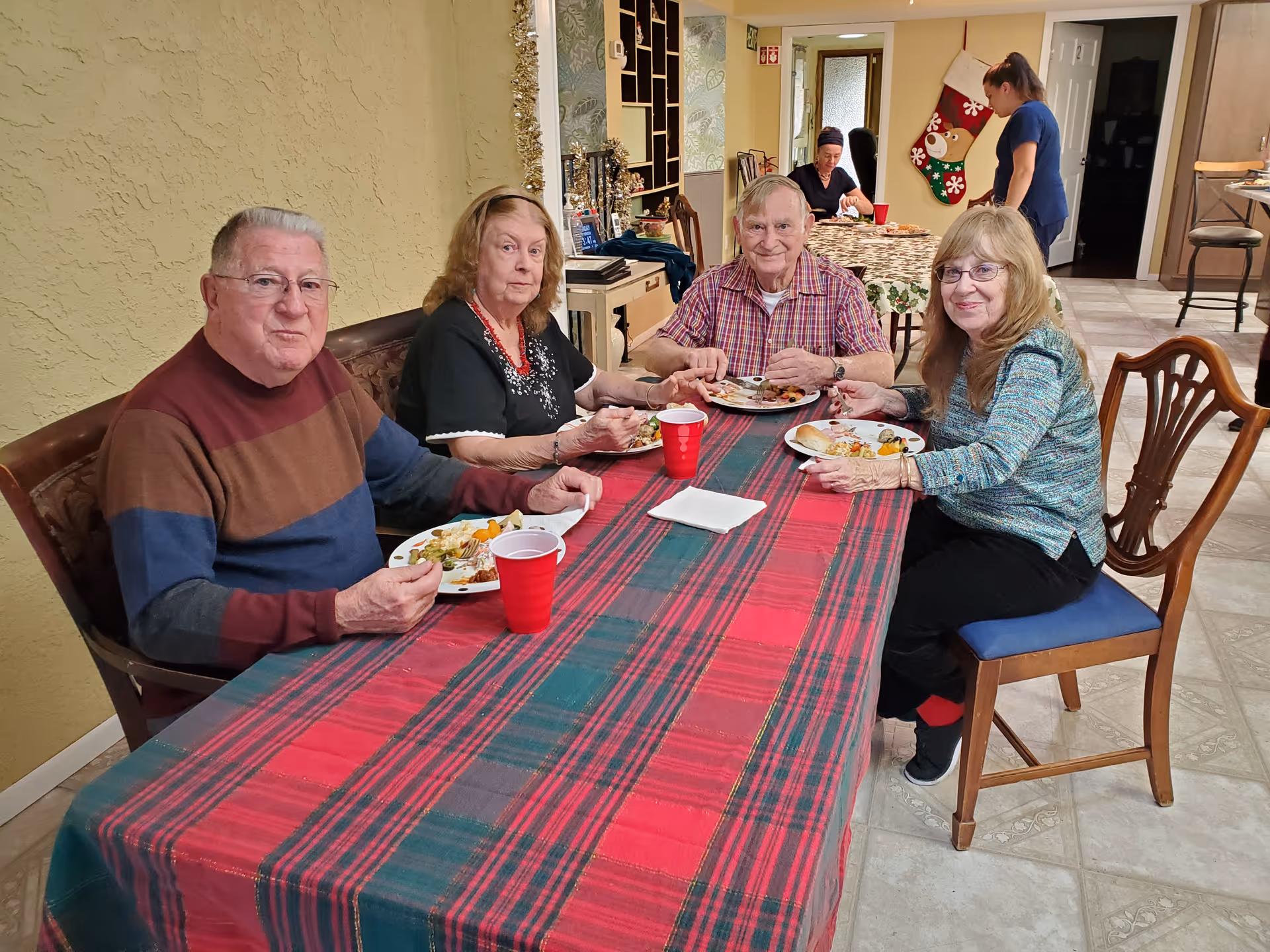 Four elderly residents sit around a holiday‑decorated dining table eating a meal in a communal dining area.