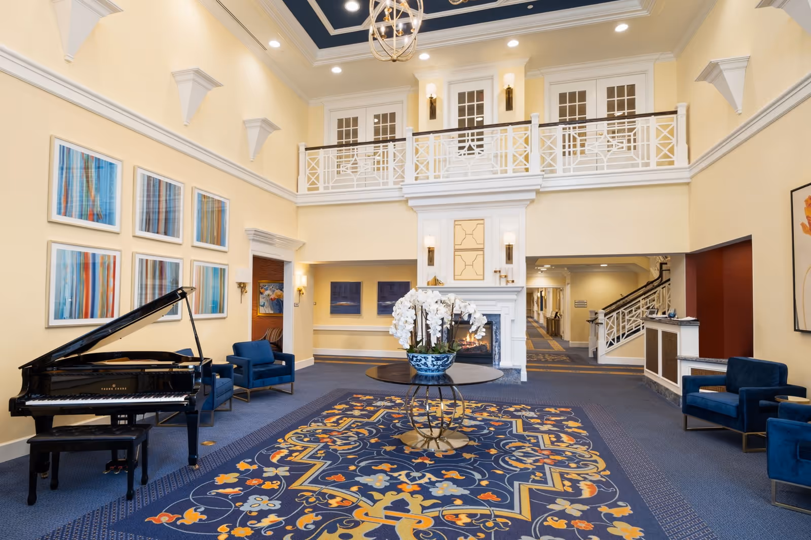 Spacious and elegant common area in an assisted living facility featuring a grand piano, blue armchairs, a large decorative rug, a round table with a floral arrangement, a fireplace, and a balcony with white railings overlooking the room.