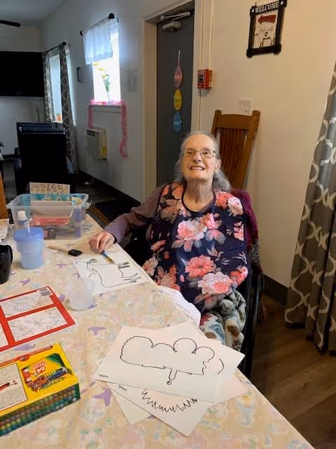 An elderly woman sitting in a wheelchair at a table covered with a pastel-colored tablecloth. She is smiling and wearing a floral bib. On the table are coloring sheets with tree outlines, crayons, a water cup, and other art supplies. The room has a window with curtains, a door with colorful hanging decorations, and a wooden chair behind her.