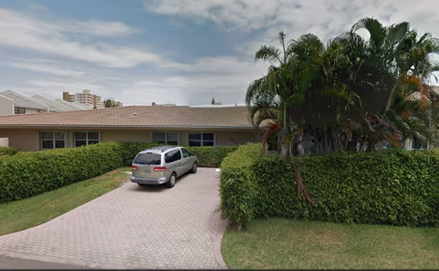 Single-story building with a tiled roof partially obscured by green hedges and palm trees. A silver minivan is parked on a paved driveway leading to the building entrance. The sky is partly cloudy.