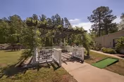Outdoor garden area at Perry County Nursing & Rehab Center featuring a wooden pergola with climbing plants, a paved walkway, green grass, trees, and a small putting green.