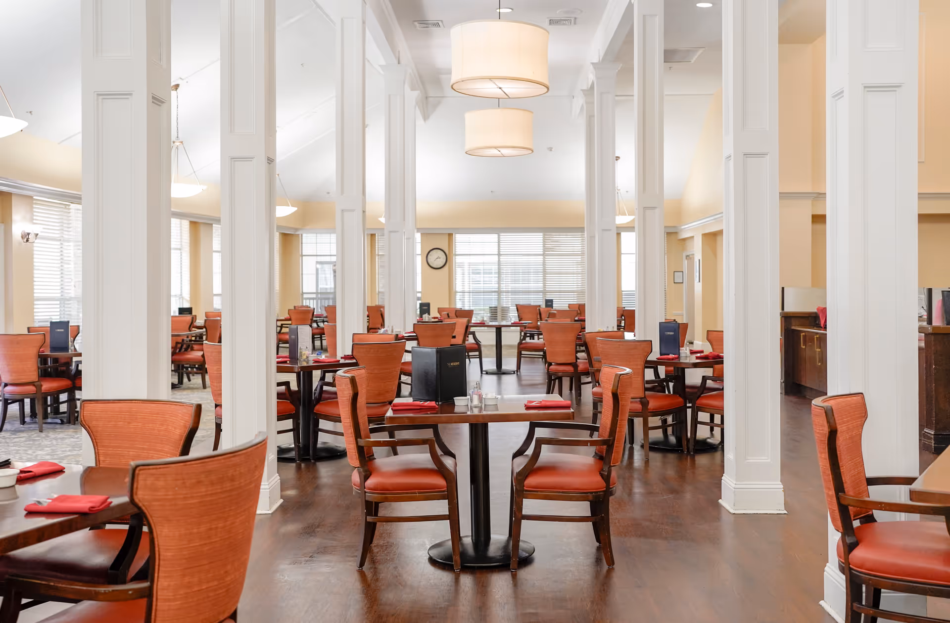 A spacious dining room with multiple wooden tables and orange cushioned chairs arranged neatly. The room features tall white columns, large windows with blinds allowing natural light, and modern hanging light fixtures. Menus and red napkins are placed on the tables.