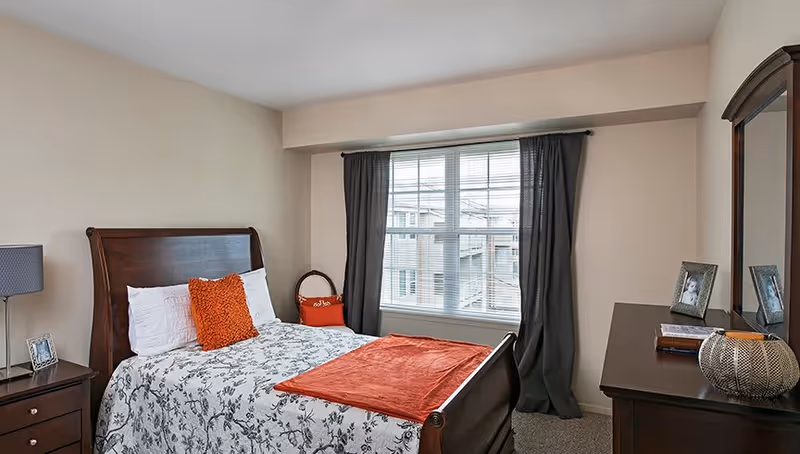 A bedroom with a wooden bed frame, a bed with white and gray floral bedding, and orange accent pillows and throw. There is a window with dark curtains, a wooden dresser with a mirror, picture frames, and decorative items. A nightstand with a lamp and a small picture frame is beside the bed.
