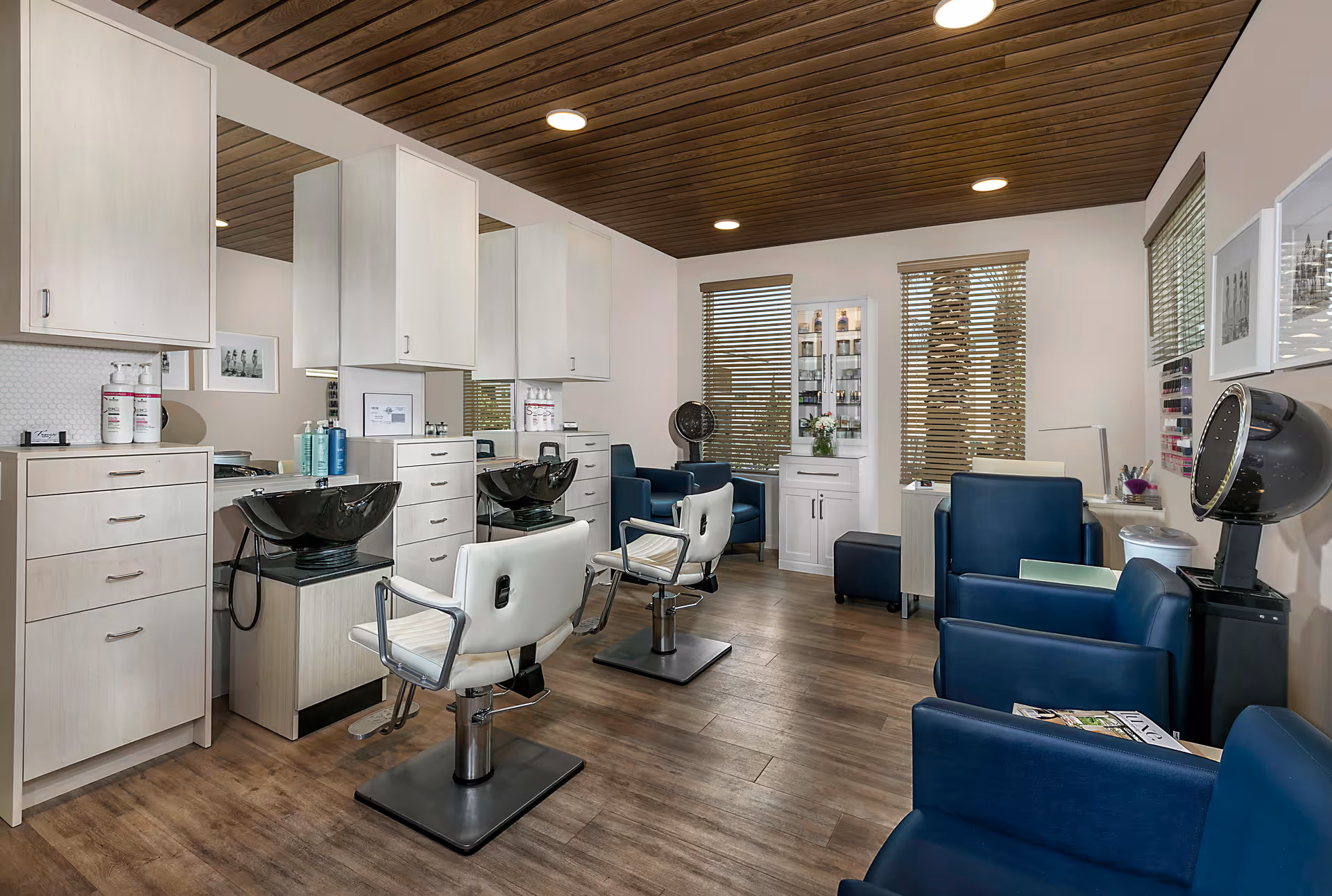 Interior of a salon room with two white salon chairs in front of black wash basins and mirrors. The room has wooden flooring, a wooden ceiling with recessed lighting, and white walls with windows covered by blinds. There are blue armchairs and salon equipment including a hair dryer and nail polish display.