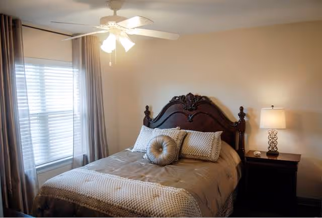 A neatly made bed with a carved wooden headboard, adorned with decorative pillows, positioned next to a window with sheer curtains. A nightstand with a lamp featuring a decorative base is beside the bed. The room has beige walls and a ceiling fan with lights.