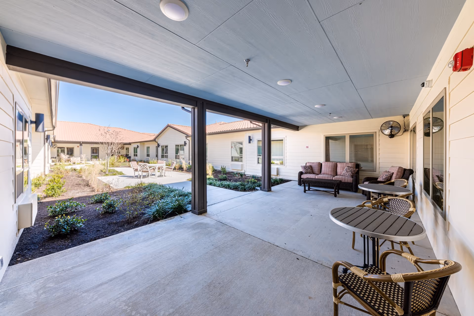 Covered outdoor patio area with round tables and chairs, a cushioned sofa and armchair, overlooking a landscaped courtyard with plants and additional seating under a clear blue sky.