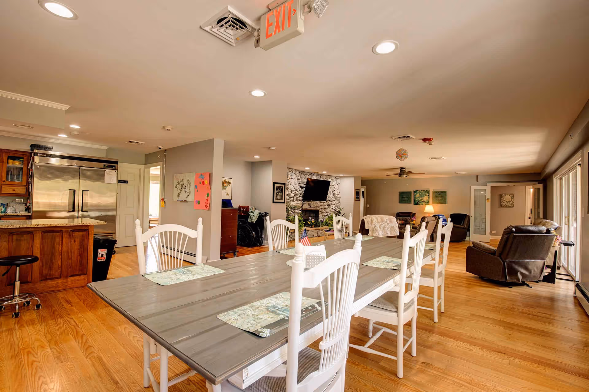 A spacious senior living common area with a long dining table and white chairs in the foreground. The room features hardwood floors, a stone fireplace with a mounted TV, several comfortable armchairs, and large windows letting in natural light. A kitchen area with a large stainless steel refrigerator is visible to the left.