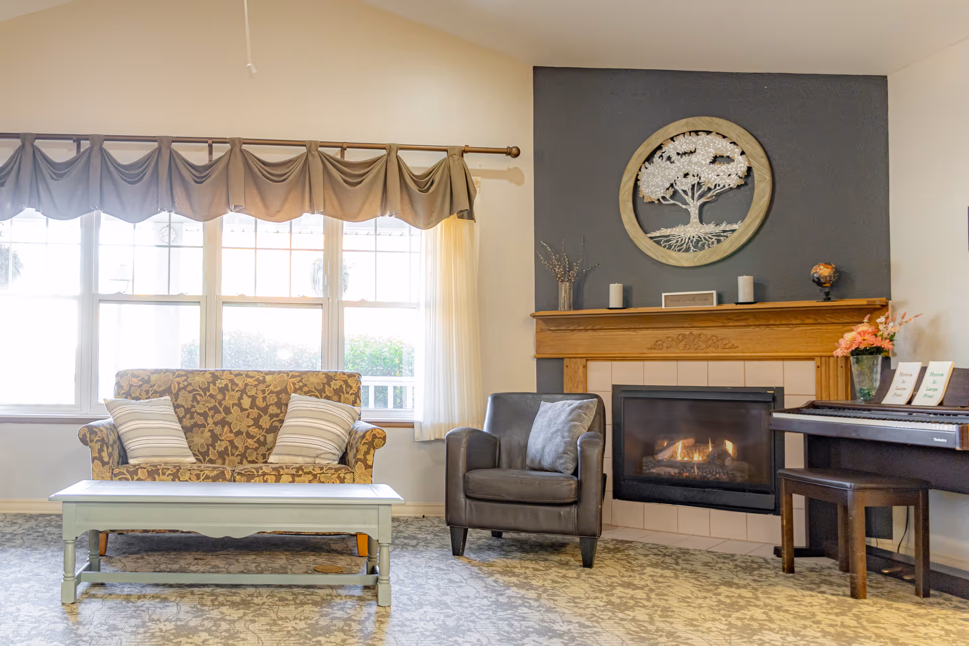 A cozy living room with a floral-patterned loveseat and two striped pillows, a gray armchair with a gray cushion, a light blue coffee table, and a fireplace with a wooden mantel. Above the mantel is a decorative round wall art featuring a tree. To the right of the fireplace is a piano with a bench and some flower arrangements on top. Large windows with beige curtains allow natural light to brighten the room.