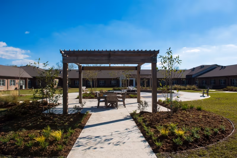 Courtyard with a wooden pergola and seating surrounded by landscaped paths and a one-story brick building under a blue sky.