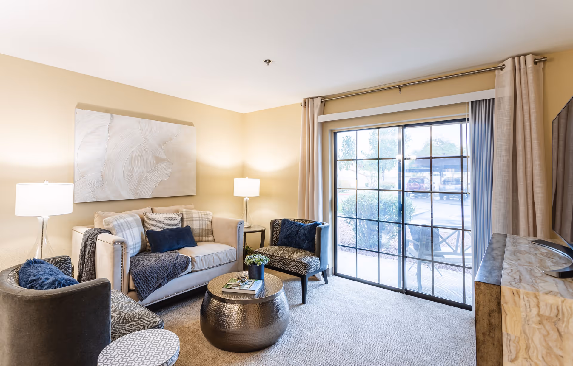A cozy living room with a beige sofa adorned with multiple cushions and a throw blanket, two patterned armchairs with dark blue pillows, a round metallic coffee table with a small plant and magazines, two table lamps on either side of the sofa, a large abstract painting on the wall, and a sliding glass door with beige curtains leading to an outdoor area.