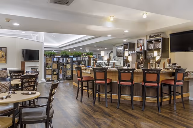 Interior view of a senior living facility lounge area with a curved bar counter and five high chairs with red cushions. The bar has various glassware and decorative items on display. To the left, there are several tables and chairs arranged for dining or socializing. The floor is wooden, and the ceiling has recessed lighting. There are plants and shelves in the background, and a flat-screen TV mounted on the wall.