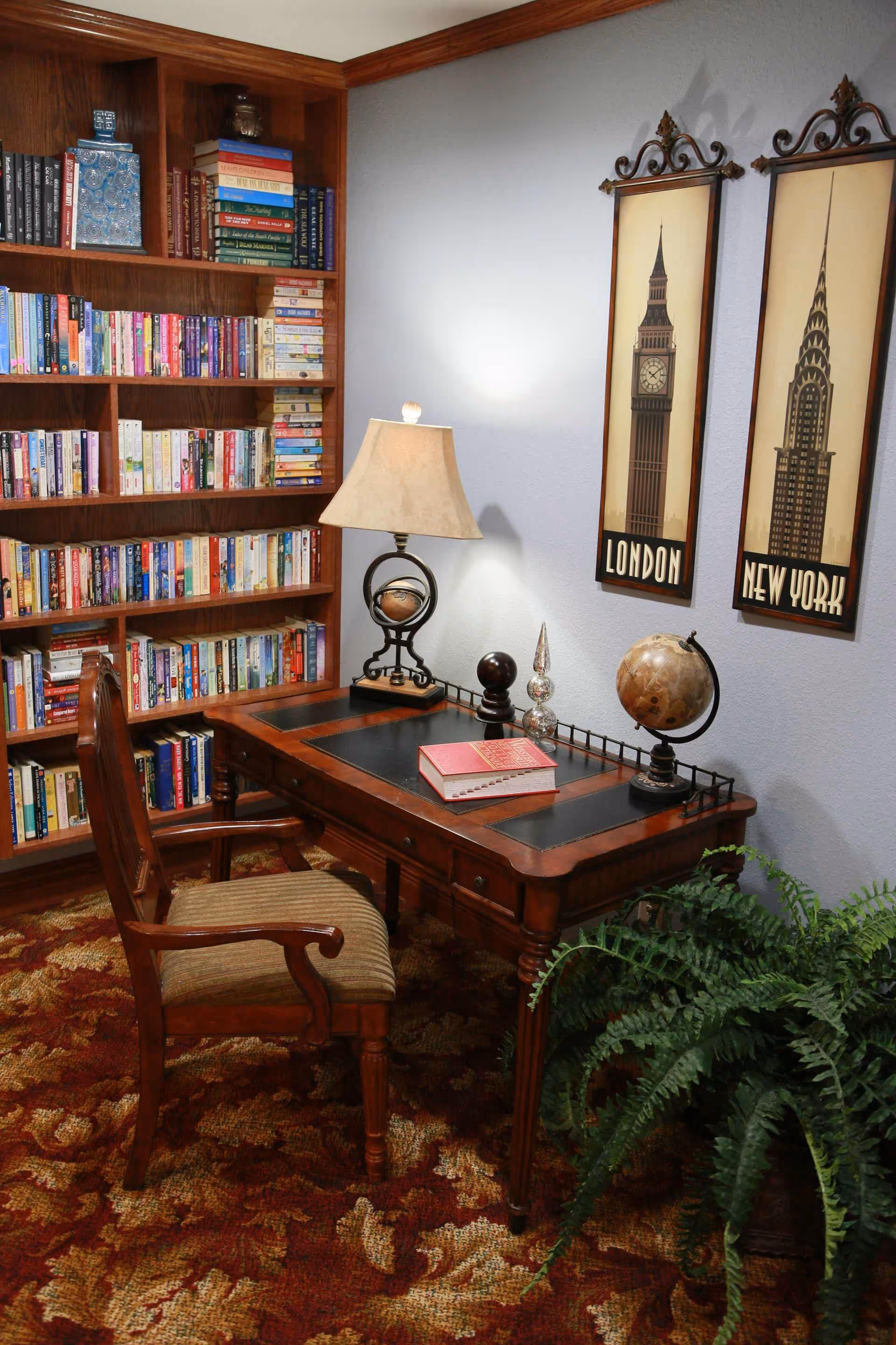 A cozy reading nook with a wooden desk and chair, lamp, globe, large bookshelf filled with books, and framed London and New York prints on the wall.