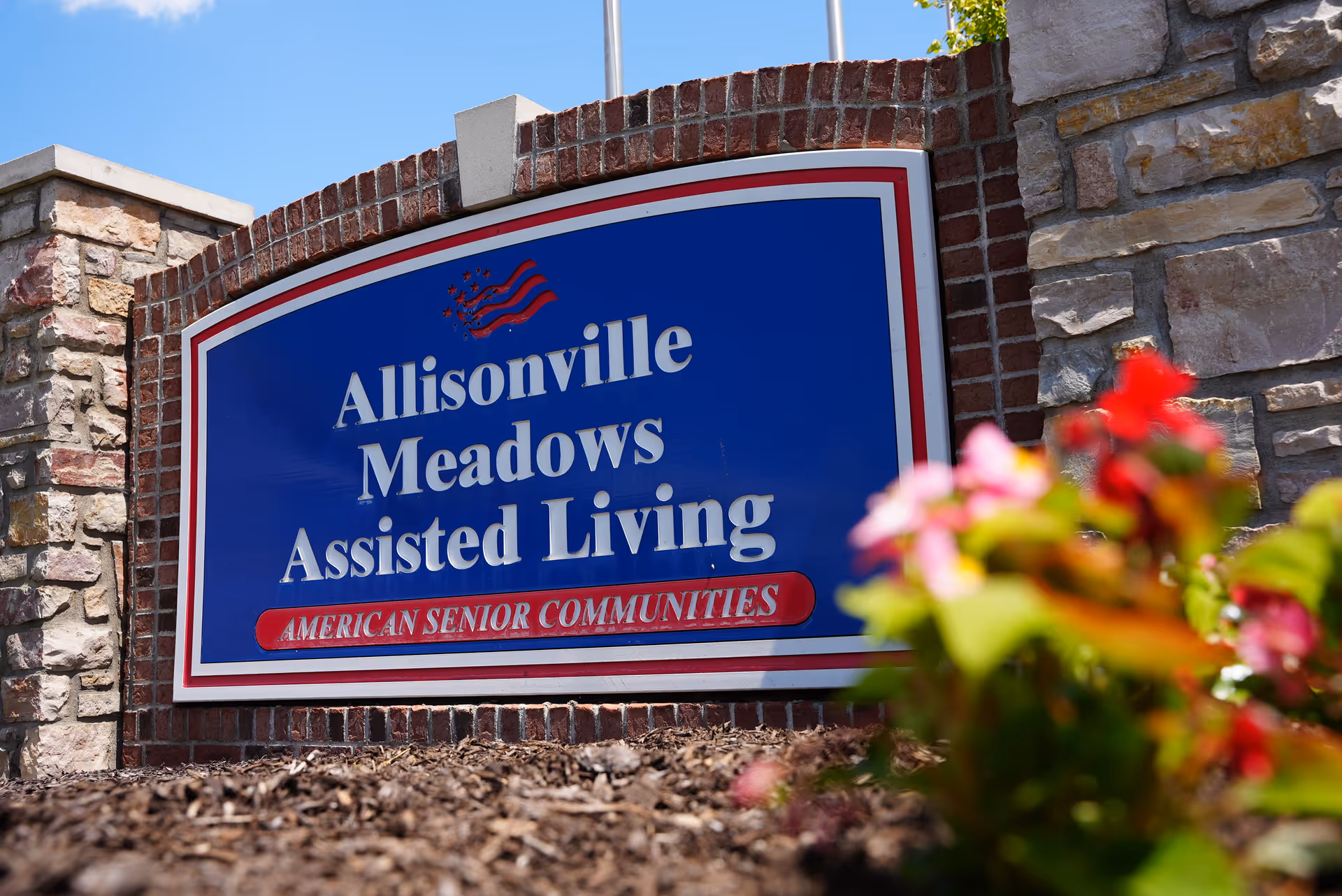 Entrance sign reading Allisonville Meadows Assisted Living mounted on a brick and stone wall with flowers in the foreground.