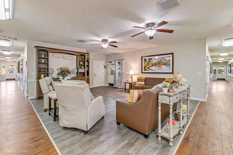 A spacious and well-lit senior living facility common area with comfortable seating including a brown leather couch and two beige recliners. The room features ceiling fans with lights, a wooden bookshelf with decorative items, a side table with a lamp, and a framed landscape painting on the wall. The area is flanked by two long hallways with wooden flooring and multiple ceiling lights.