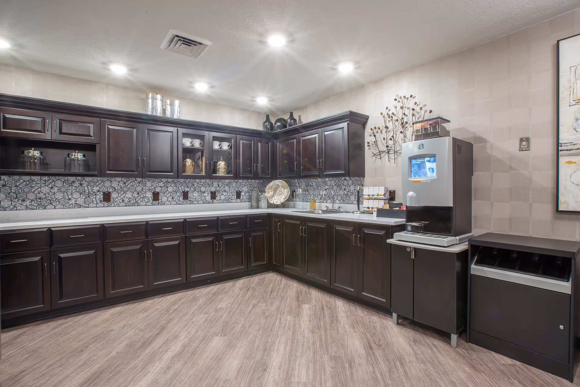 A clean and modern kitchen area with dark wooden cabinets, a patterned tile backsplash, and a light-colored countertop. There is a coffee machine on a small counter to the right, along with a trash bin. The floor is a light wood laminate, and the ceiling has recessed lighting.