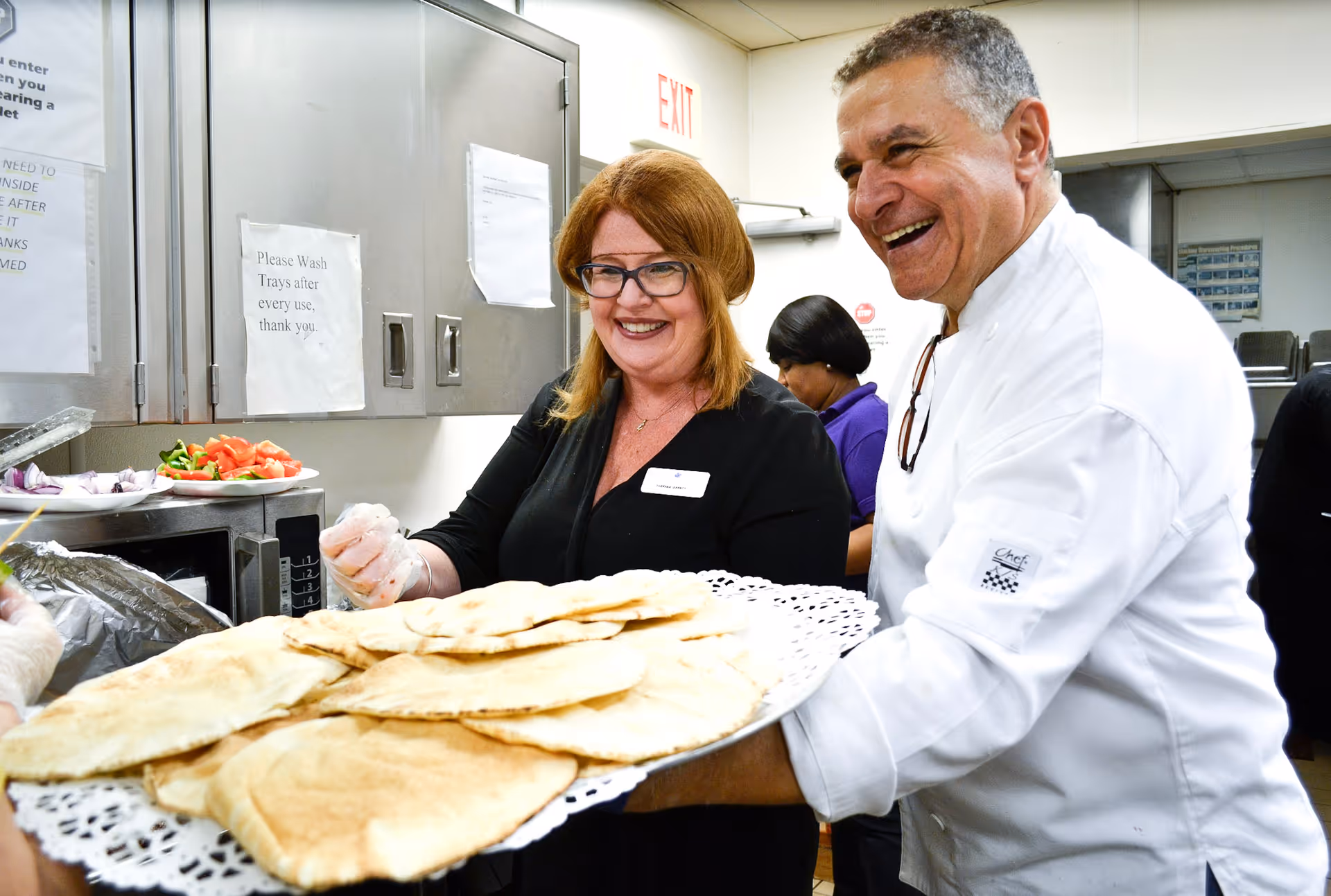 Two smiling staff members in a commercial kitchen holding a large tray of pita bread.