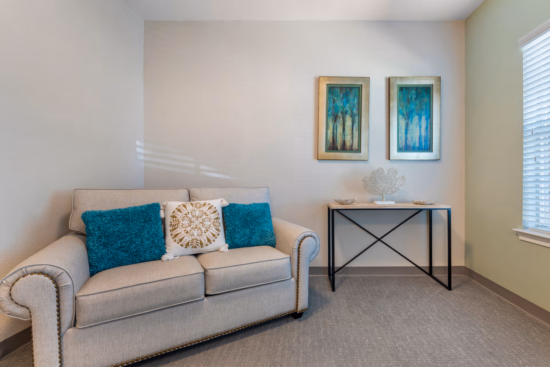 Small furnished living room with a beige sofa topped with teal and patterned pillows, a narrow console table with decorations, two framed paintings, and a window with blinds.
