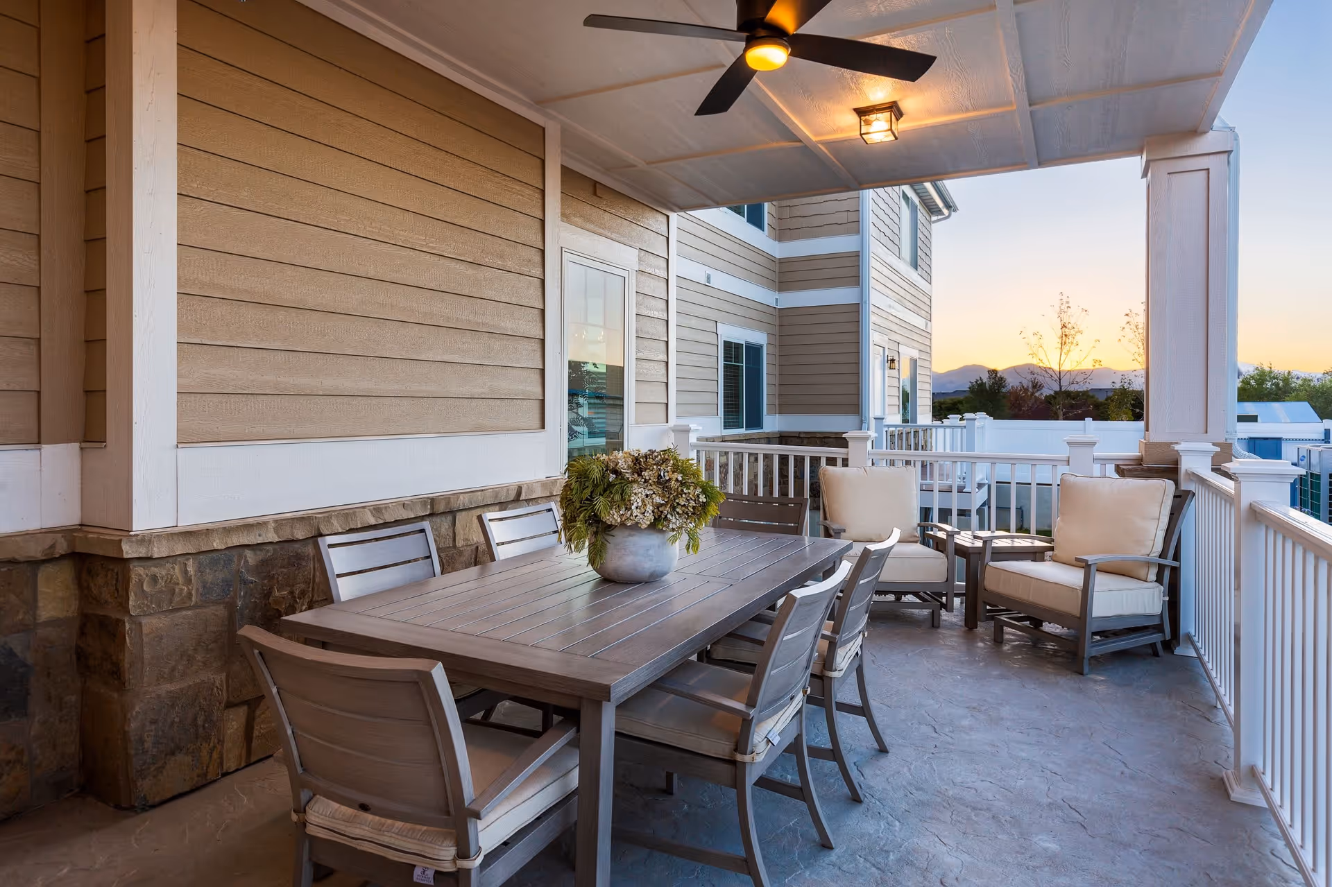 Covered outdoor patio with a dining table, chairs, and cushioned lounge seating overlooking nearby buildings at sunset.
