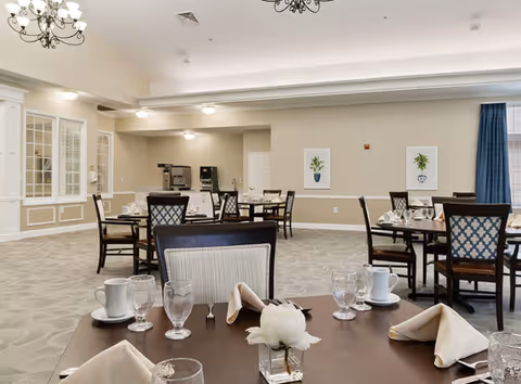 A spacious dining room with several tables set with white napkins, glassware, and cups. The room has beige walls, carpeted floors, and a coffee station in the back corner. There are decorative framed pictures on the walls and a chandelier hanging from the ceiling.