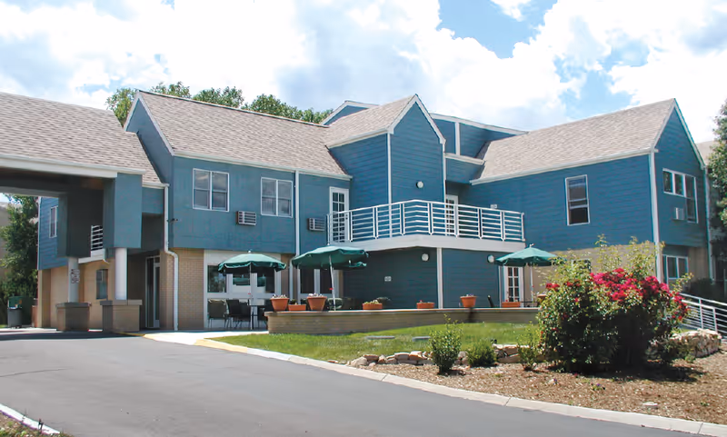 Exterior view of a two-story assisted living facility building painted blue with white trim. The building features a balcony with white railings, several windows, and a covered entrance area. There are green patio umbrellas and outdoor seating on a paved area near the entrance. Landscaping includes bushes, flowers, and a small garden bed with rocks. The sky is partly cloudy.