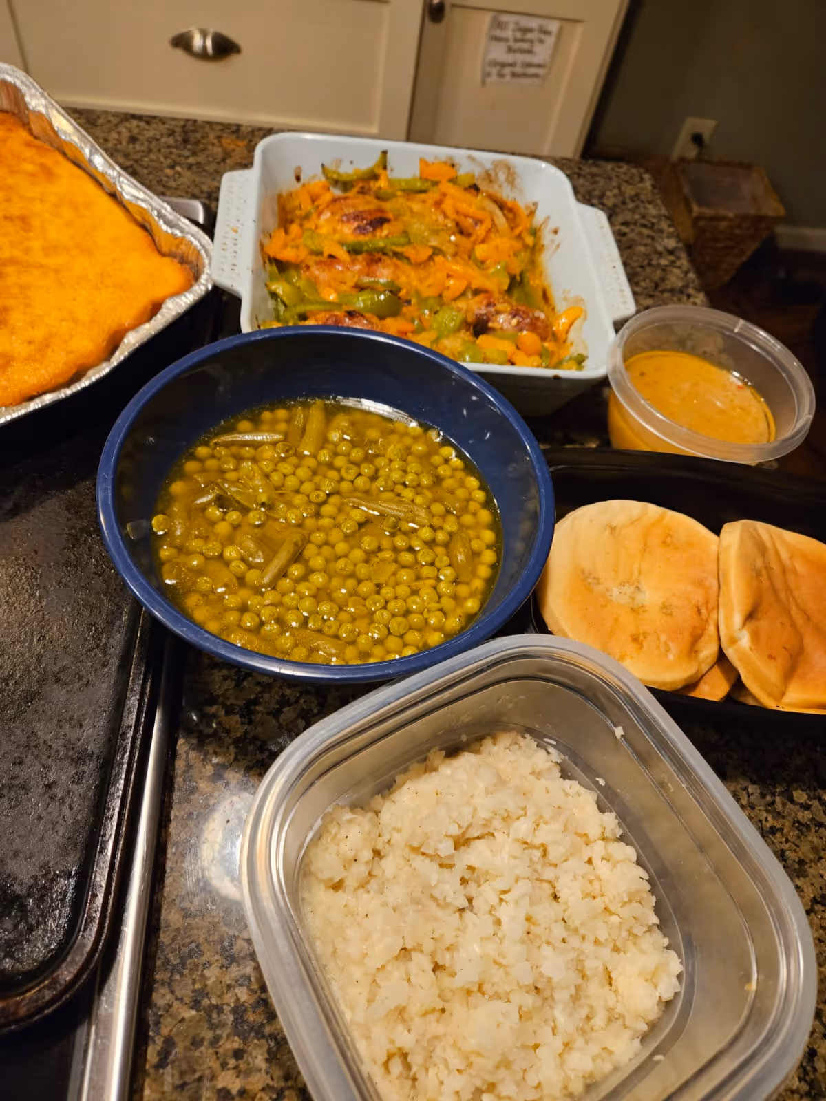 Countertop with containers of prepared food including a bowl of peas, a container of rice, roasted vegetables, cornbread and rolls.