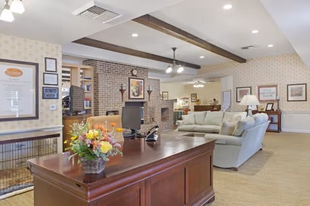 A spacious and well-lit living room area in a senior living facility featuring a wooden desk with a flower arrangement, a computer, and a phone. Behind the desk, there is a cozy seating area with light-colored sofas, a brick fireplace, framed artwork on the walls, and wooden ceiling beams. The room has a warm and inviting atmosphere with soft lighting and patterned wallpaper.