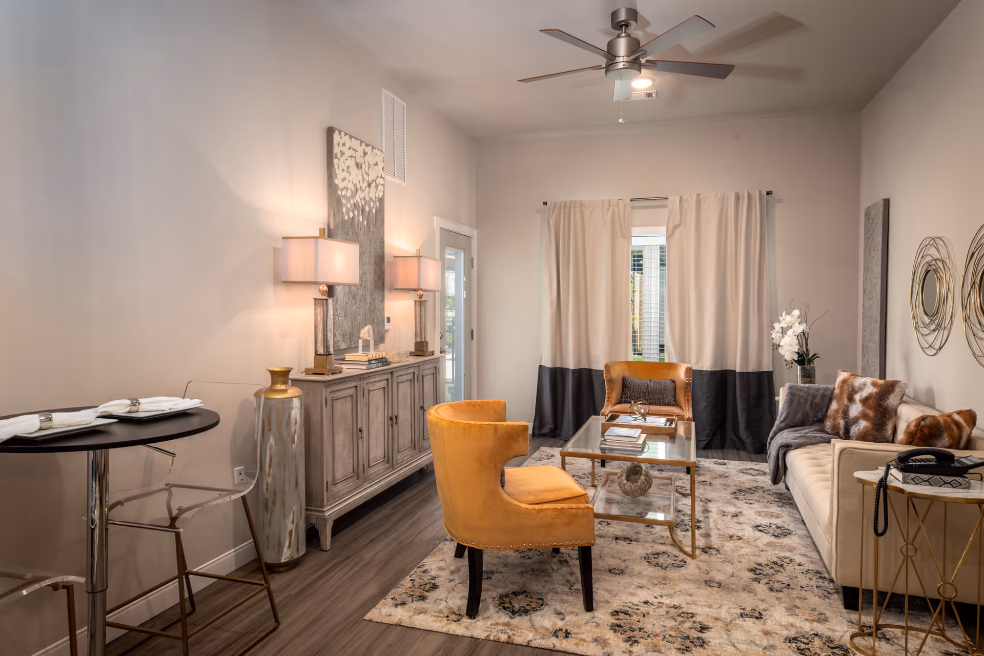Well-appointed living room with a beige sofa, two mustard accent chairs, a glass coffee table, sideboard with lamps, and a patterned rug.