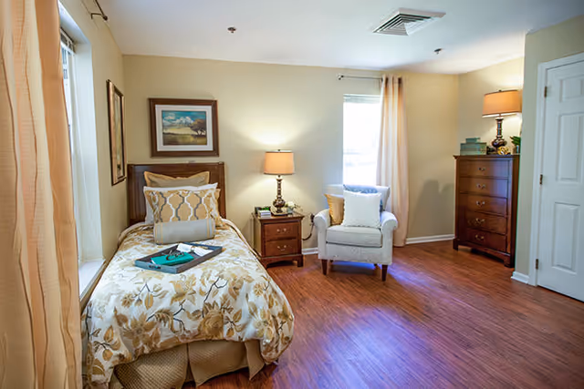 Well-lit bedroom with a single bed, nightstand and lamp, upholstered armchair, and tall dresser on wood flooring.