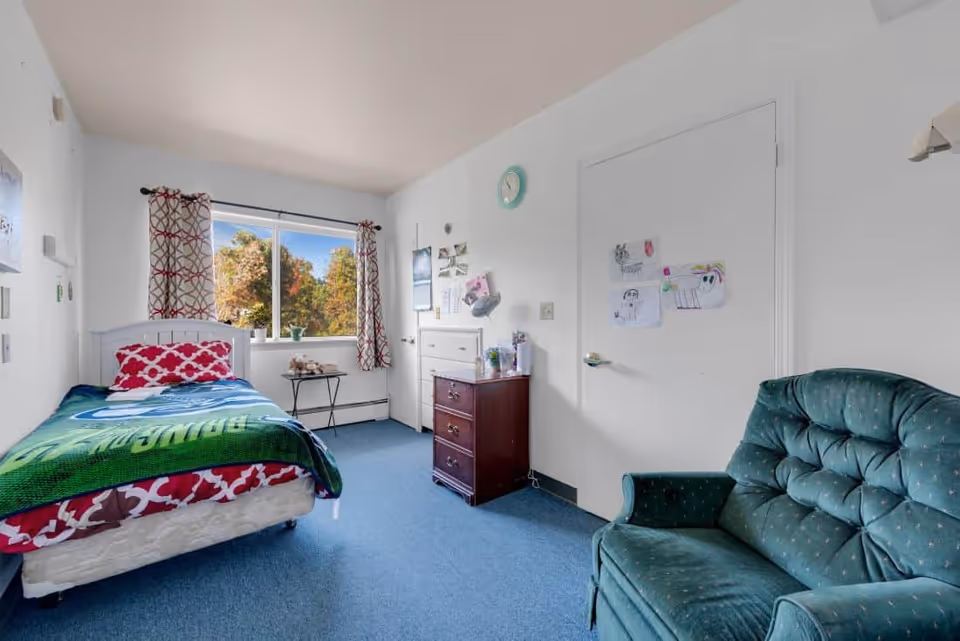 A small bedroom with a single bed covered with a green and red blanket, a red patterned pillow, a window with patterned curtains showing trees outside, a wooden dresser with items on top, a green upholstered armchair, and children's drawings taped to a white door.