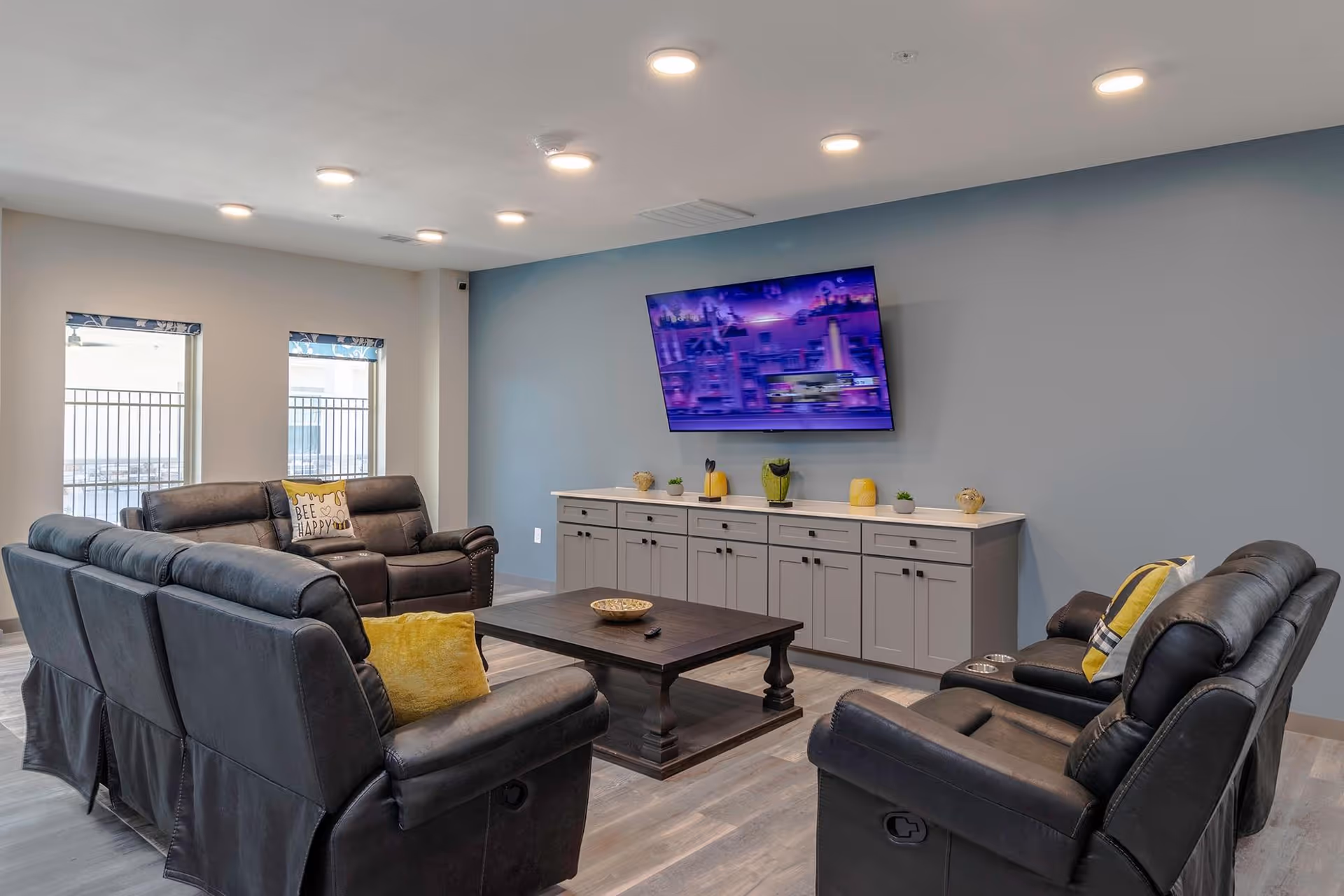 A modern living room with black leather recliner sofas arranged around a dark wooden coffee table. A large flat-screen TV is mounted on a light blue accent wall above a long gray cabinet with decorative items. Two windows with blinds allow natural light into the room.