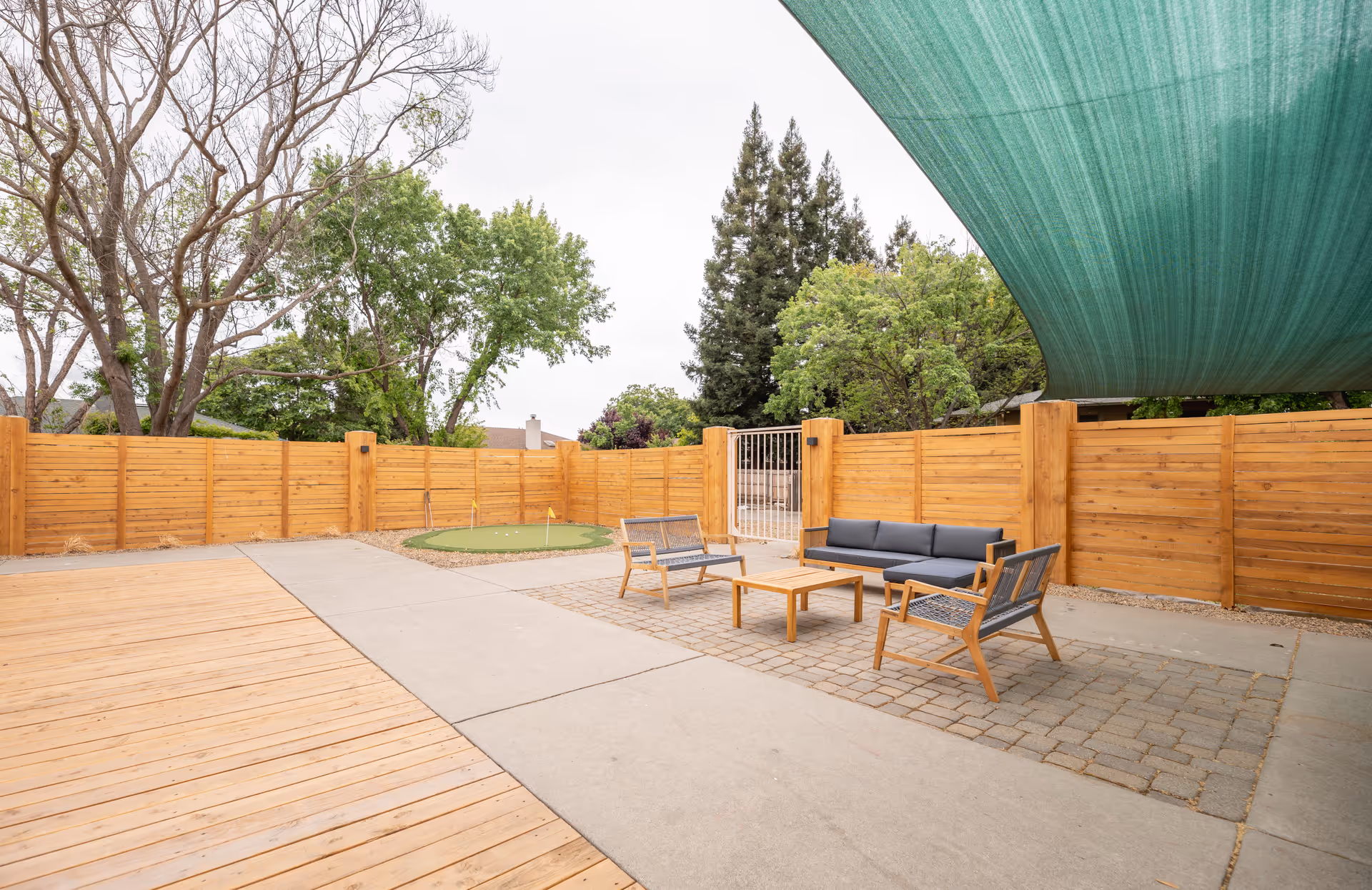 Outdoor patio area with wooden fencing, a green shade canopy overhead, and outdoor seating including a sofa, two chairs, and a coffee table. There is also a small putting green in the background surrounded by trees.