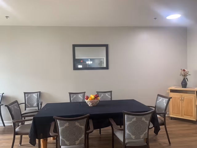 A dining area with a rectangular table covered with a black tablecloth, surrounded by eight chairs with patterned upholstery. A basket of fruit is placed in the center of the table. On the right side, there is a wooden cabinet with a vase of flowers on top. A rectangular mirror hangs on the beige wall above the table.