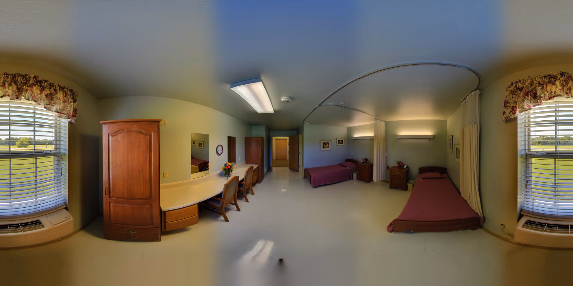 Wide-angle view of a shared senior care bedroom with two single beds, desks, chairs, a wardrobe, and windows.