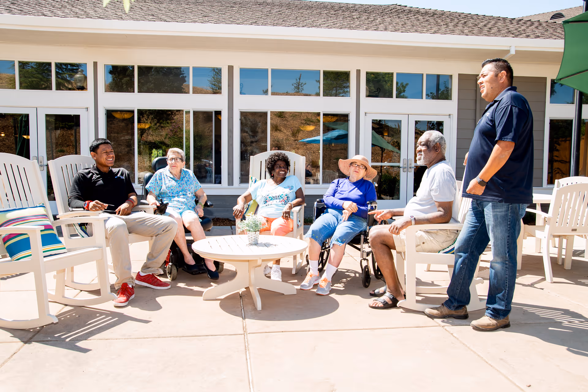 A group of five seniors and one staff member sitting and standing around a round outdoor table on a sunny patio at TreVista Senior Living. They are engaged in conversation and appear relaxed and happy. The patio has white Adirondack chairs and large windows of the building are visible in the background.