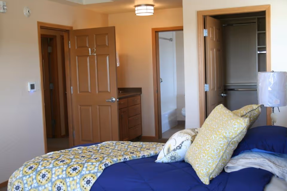 Interior view of a bedroom in an assisted living facility showing a bed with blue and yellow patterned bedding and pillows. The room has beige walls and wooden trim. There are three open doors visible: one leading to a bathroom with a bathtub and toilet, one to a closet, and one to a hallway. A lamp with a white shade is on a bedside table.