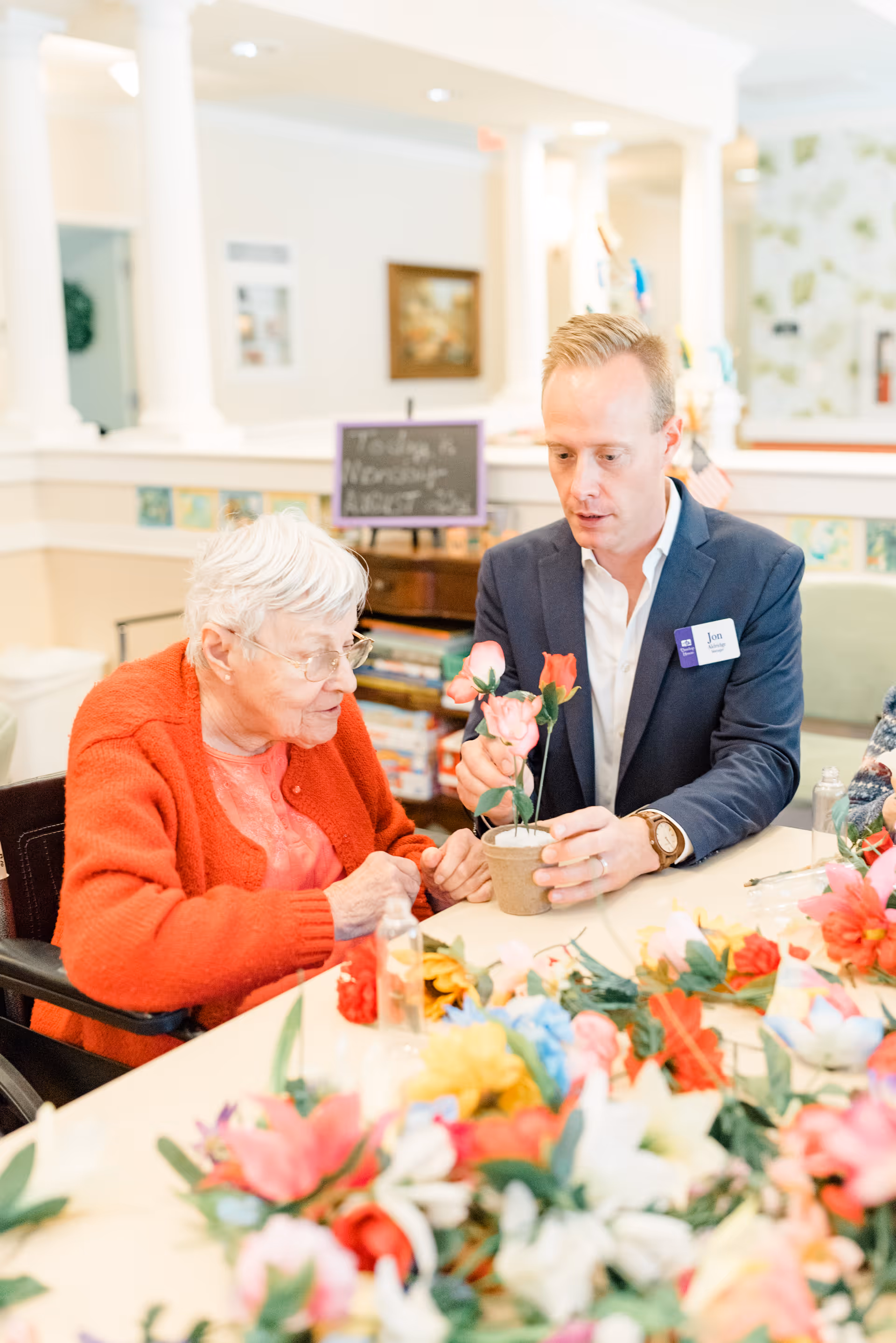 A staff member helps an elderly woman arrange flowers at a table in a bright assisted living common area.