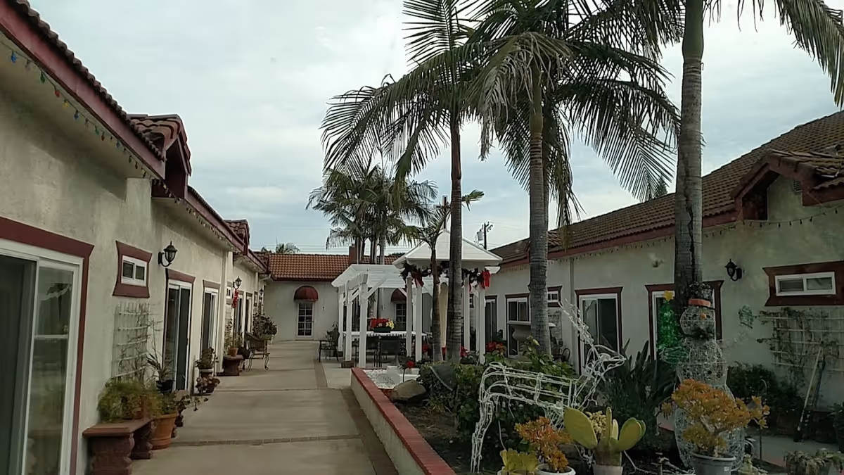 Outdoor courtyard area of Cardinal Yu Pin Manor featuring a walkway lined with potted plants and palm trees. There is a white pergola structure decorated with holiday wreaths and a garden area with wireframe holiday decorations including a reindeer and a snowman. The buildings surrounding the courtyard have beige walls with red trim and tiled roofs.