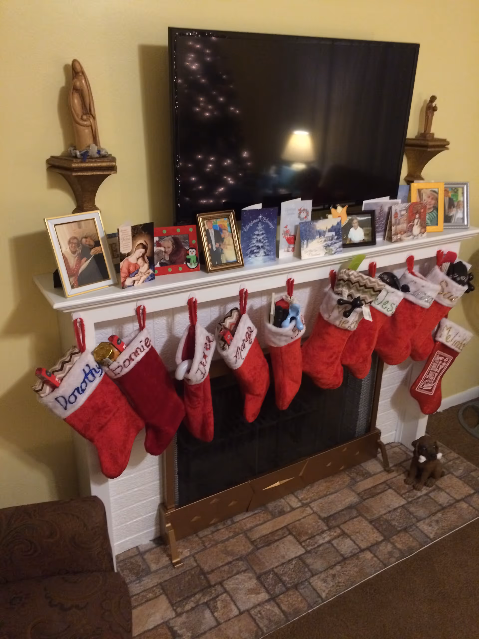 A living room fireplace mantel decorated with Christmas stockings, holiday cards, framed photos, and a TV mounted above.