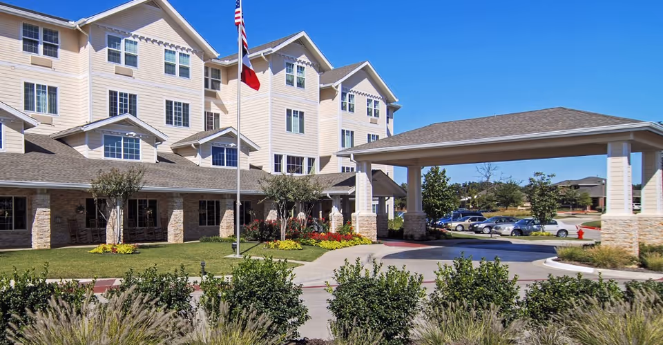 Exterior view of Dogwood Estates - Sky Active Living facility showing a multi-story building with beige siding and stone accents, a covered entrance driveway, an American flag and Texas state flag on a flagpole, landscaped greenery, and parked cars in the background under a clear blue sky.