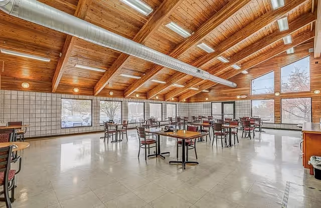 Spacious dining room with multiple tables and chairs arranged neatly. The room features a high wooden ceiling with exposed beams and large windows allowing natural light to fill the space. The floor is tiled, and there is a visible ventilation duct running along the ceiling.